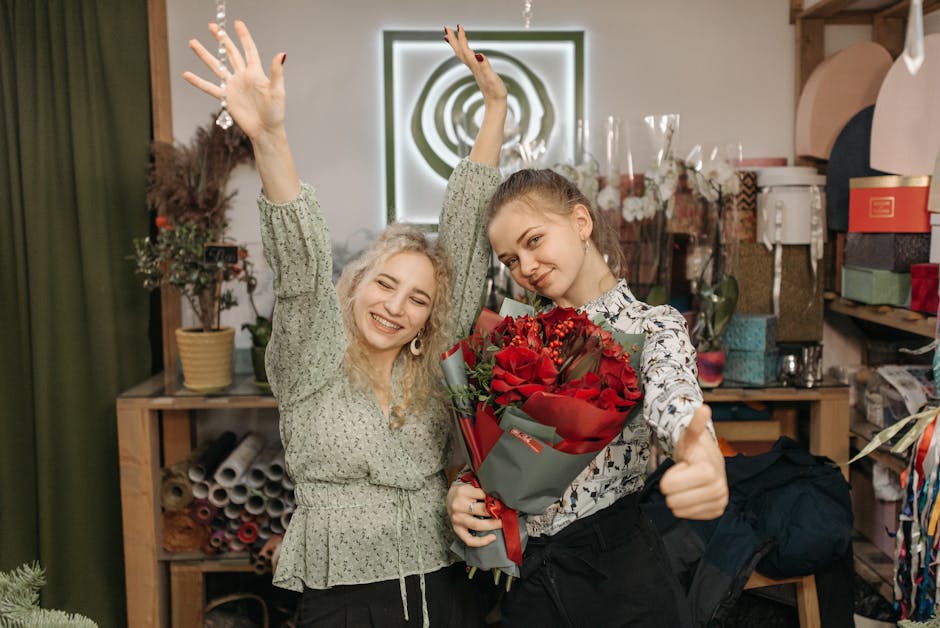 Two women inside a florist shop are celebrating with smiles and gestures of joy, one holding a bouquet of vibrant red roses wrapped in grey paper with red ribbon, and the other with her hands raised i