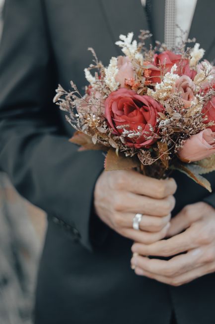 A close-up of a person dressed in a dark suit holding a floral bouquet, which features large pink roses accompanied by smaller white and beige filler flowers, all arranged in a rounded, compact style.